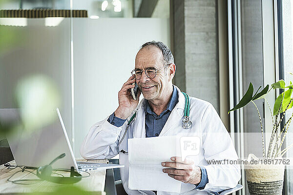 Smiling mature doctor with medical report talking on smart phone at desk