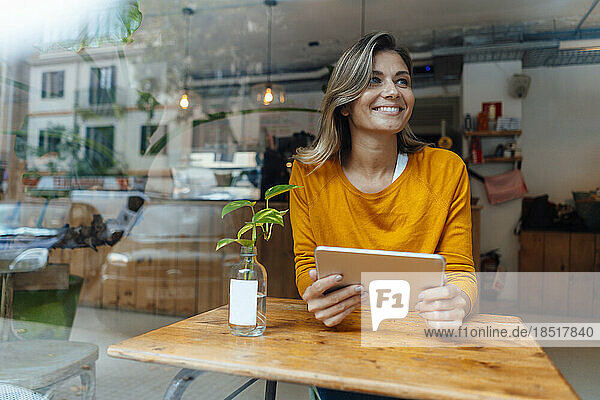 Happy woman sitting with tablet computer in cafe seen through glass