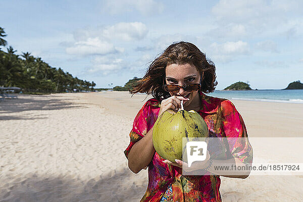 Lächelnde junge Frau trinkt Kokosnusswasser am Strand