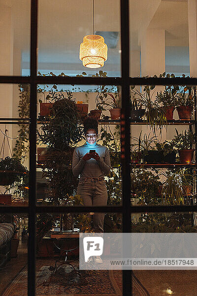 Businesswoman using mobile phone standing by plants at night seen through glass