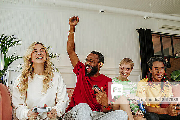Excited young man with hand raised sitting by friends playing video game at home