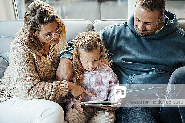 Daughter using tablet PC sitting with parents on sofa in living room