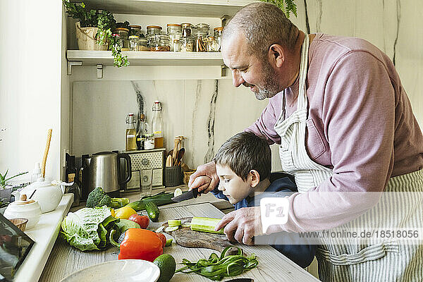 Grandfather teaching grandson to cut vegetables in kitchen at home