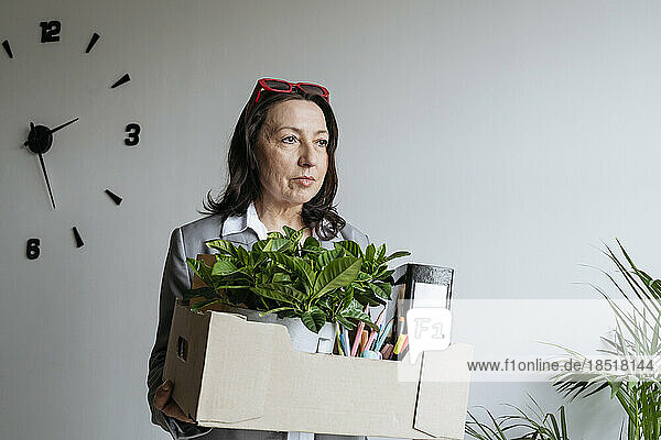 Sad businesswoman holding box with office supplies in front of wall