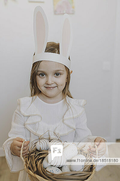 Smiling girl wearing rabbit ears holding basket of Easter eggs at home