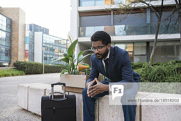 Deprimierter Geschäftsmann mit Koffer und seinen Habseligkeiten sitzt vor Bürogebäude