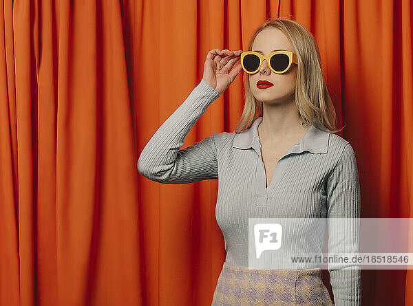 Blond woman wearing sunglasses standing in front of orange curtain