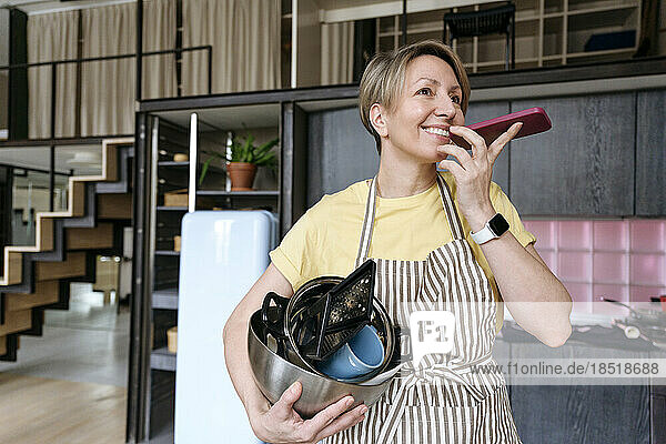Frau hält Geschirr in der Hand und spricht zu Hause mit dem Smartphone