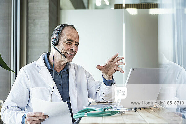 Smiling mature doctor explaining over telemedicine video call at desk