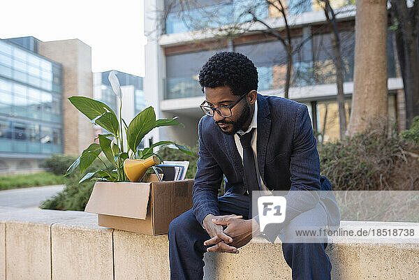 Gestresster Geschäftsmann mit Karton sitzt an der Wand in der Nähe eines Bürogebäudes