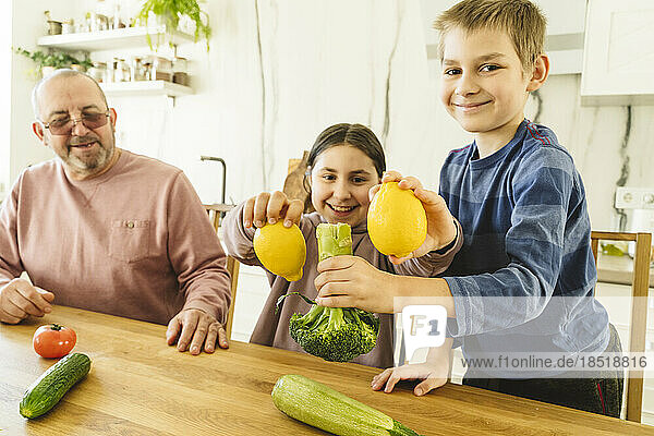 Glücklicher Großvater mit Enkel und Enkelin  die Gemüse am Esstisch halten