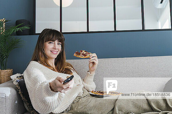 Lächelnde Frau hält Pizza in der Hand und schaut zu Hause auf dem Sofa fern