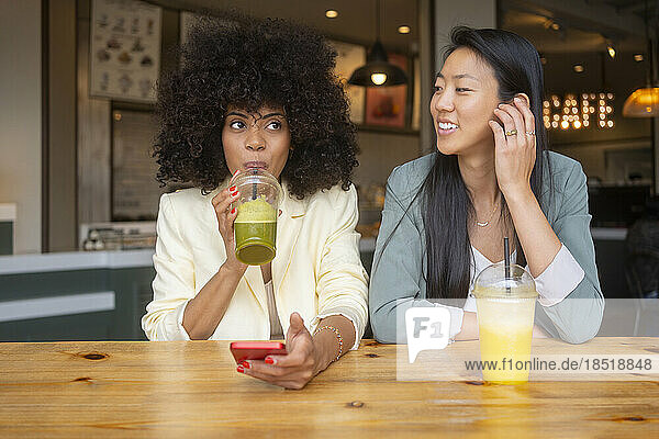 Smiling woman looking at friend drinking juice in cafe