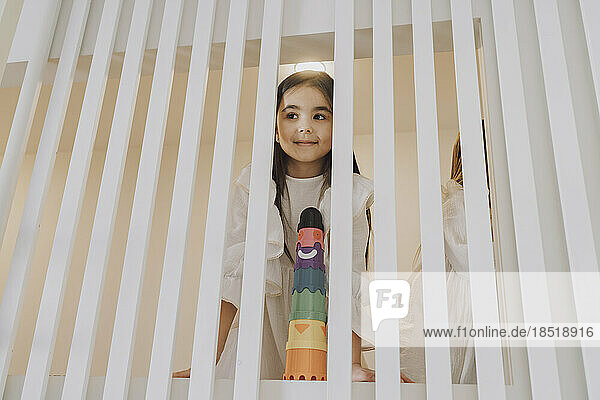 Girl with toy looking through striped railing at home