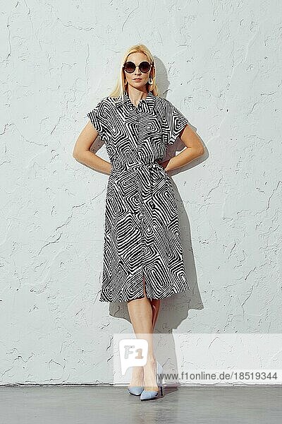 Relaxed blonde woman in a patterned dress and sunglasses leaning against white wall in the studio