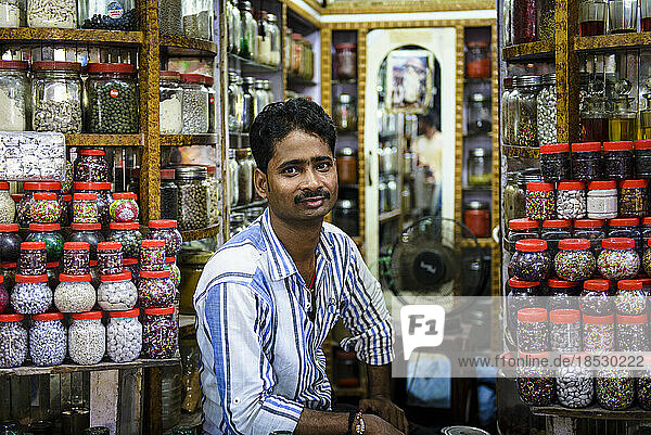 Männlicher Ladenbesitzer sitzt in seinem Süßwarenladen voller Gläser mit Süßigkeiten; Varanasi  Indien