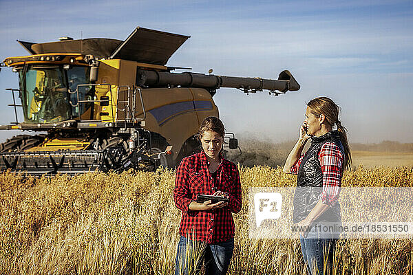 A mature farm woman standing in a field making a call and working together with a young woman at harvest time  using advanced agricultural software on a pad  while a combine harvester works in the background; Alcomdale  Alberta  Canada