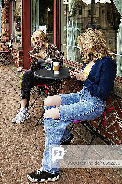 Zwei Teenager genießen die Zeit miteinander und sitzen an einem Tisch für einen Kaffee in einem Café im Freien und verbringen Zeit mit ihren Smartphones; St. Albert  Alberta  Kanada
