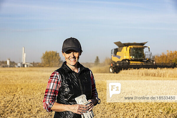 Porträt einer reifen Bäuerin  die während der Ernte in einem Getreidefeld steht und für die Kamera posiert  mit einem arbeitenden Mähdrescher und einer petrochemischen Anlage im Hintergrund bei Sonnenuntergang; Alcomdale  Alberta  Kanada