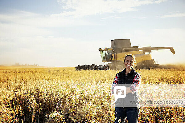 Porträt einer reifen Bäuerin  die während der Ernte in einem Getreidefeld steht und für die Kamera posiert  während im Hintergrund bei Sonnenuntergang ein Mähdrescher arbeitet; Alcomdale  Alberta  Kanada
