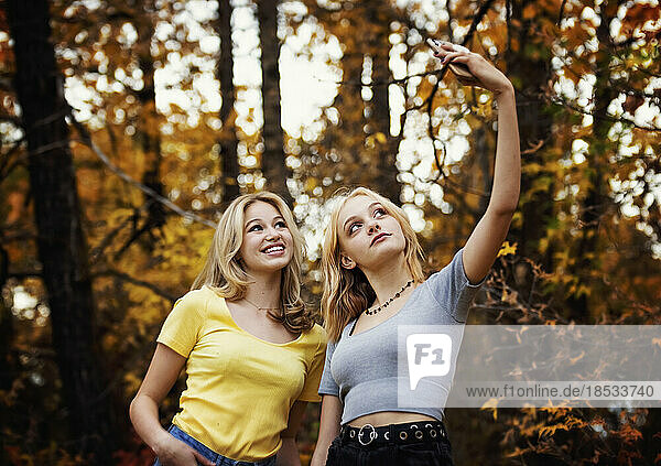 Zwei Mädchen im Teenageralter machen Selbstporträts mit einem Smartphone  während sie an einem warmen Herbsttag in einem Stadtpark Zeit miteinander verbringen; St. Albert  Alberta  Kanada.
