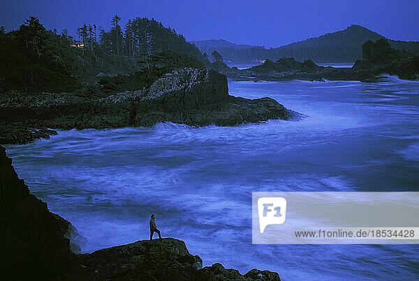 Lone figure watches the surf pound a rocky coast as night falls; Vancouver Island  British Columbia  Canada