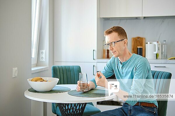 Middle aged man is sitting behind the kitchen table and holding a pill and a glass of water in hands