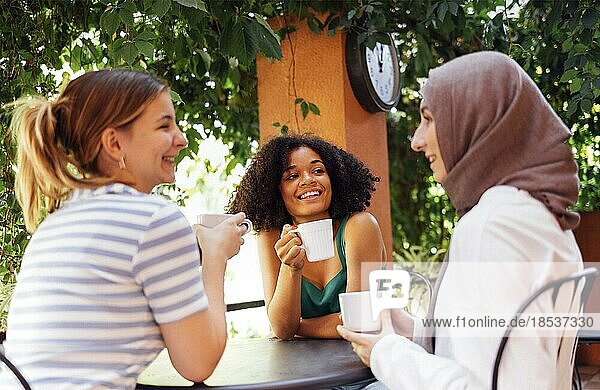 Multiethnische Gruppe von Mädchen in Freizeitkleidung und traditionellem Hijab  die sich im Freien unterhalten und Spaß haben. Drei junge Teenagermädchen in einem Gartencafé trinken Tee oder Kaffee