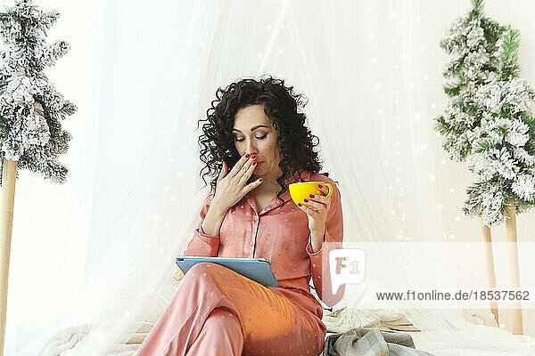 Junge attraktive  überraschte Frau im Pyjama mit einer Tasse Morgenkaffee  die auf einen digitalen Tablet Bildschirm schaut  einen erstaunten Gesichtsausdruck macht und den Mund mit der Hand bedeckt  auf einem Himmelbett zu Hause sitzend