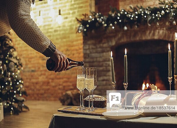 Silvesterfeier. Mann gießt Champagner in Gläser stehen auf dem Tisch mit festlichen Weihnachtsessen  Kerzen und eingepackt Geschenke gegen unscharfen Hintergrund von Backstein Kamin und firtree  beschnitten