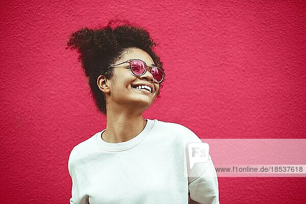 Junge Frau afrikanischer Abstammung mit modischer Sonnenbrille und lockigem Haar  das zu einem hohen Pferdeschwanz gebunden ist. Sie schaut weg und lächelt breit  während sie gerade  perfekte Zähne zeigt und vor einem Wandhintergrund posiert