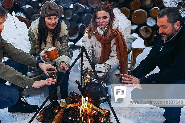 Eine Gruppe von Freunden versammelt sich um ein Lagerfeuer im Hinterhof  trinkt Tee und wärmt sich die Hände. Zwei glückliche Paare  die sich entspannen und die Wintersaison genießen  während sie am Feuer sitzen. Outdoor Winter unterhaltsam