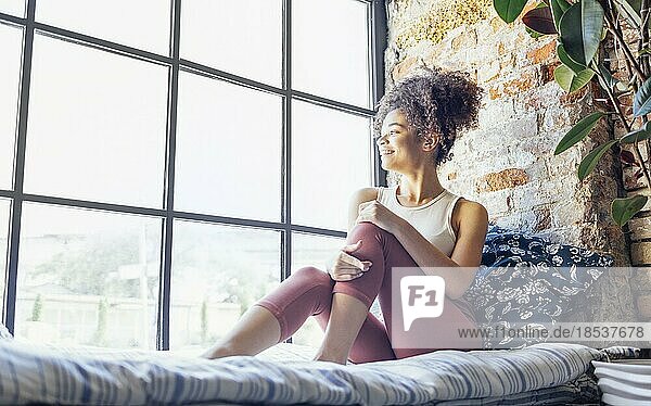 Pleased relaxed African American ethnicity girl in homewear sitting on windowsill  happy afro female at home smiling gently looking to the window
