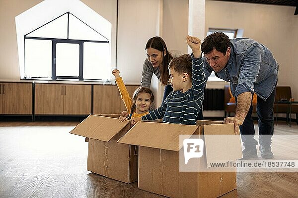 Glückliche Eltern mit Kindern im Wohnzimmer  zwei niedliche Kinder  Junge und Mädchen  sitzen lachend in Kartons und schieben Mutter und Vater. Die Familie feiert den Umzugstag