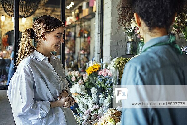 Glückliche schwarze Unternehmerin steht in einem Blumenladen und verkauft frische Blumen an Kunden. Junge blonde Mädchen kaufen einen frischen Blumenstrauß von Florist. Lächelnde afrikanische Frau Botaniker  Verkauf von Blumen