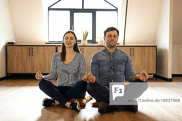 Friedliches erwachsenes Paar in Sportkleidung sitzt auf dem Boden im Lotussitz mit geschlossenen Augen und meditiert zusammen zu Hause. Ruhige Menschen  Mann und Frau  mit Yogapraxis Entspannung Konzept