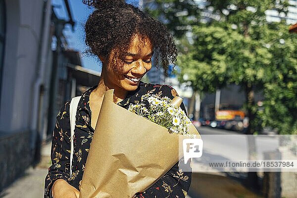 Junge afroamerikanische Frau hält Blumen und lächelt. Glückliche schöne dunkelhäutige Mädchen geht entlang der Straße mit einem Blumenstrauß von Gänseblümchen. Positive Modell im Freien auf der Stadt Sommer Hintergrund. Kopieren Raum
