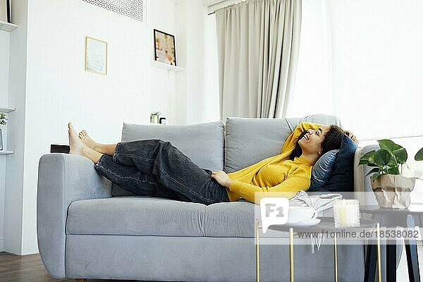 Gelassene  entspannte junge afroamerikanische Frau  die wegschaut  auf dem Sofa liegend  ruhig und lächelnd  die sich zu Hause ausruht und das faule Wochenende zu Hause genießt