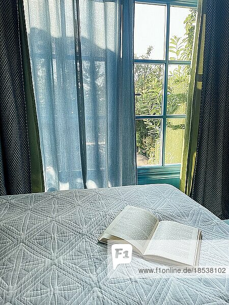 Ein aufgeschlagenes Buch liegt auf einer weißen Steppdecke auf dem Bett. Das Sonnenlicht scheint und der grüne Garten ist durch die Schlafzimmerfenster sichtbar. Gemütliches Interieur. Zeit für sommerliche Ruhe und Entspannung