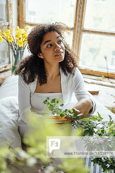 Hübsches romantisches afroamerikanisches Mädchen  das auf der Fensterbank sitzend ein Buch liest und mit Blumen überhäuft wird. Schöne Afro Frau liest fröhlich einen Roman in gemütlichem Zimmer. Bildung Konzept