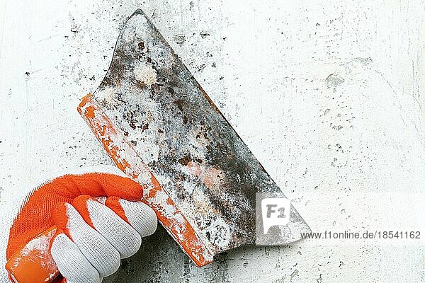 Unkenntlich gemachte Hand eines Stuckateurs hält ein altes Spezialspachtel und kratzt eine Betoninnenwand ab  entfernt alten Putz bei der Renovierung eines Gebäudes. Nahaufnahme  selektiver Fokus auf Arbeitswerkzeug
