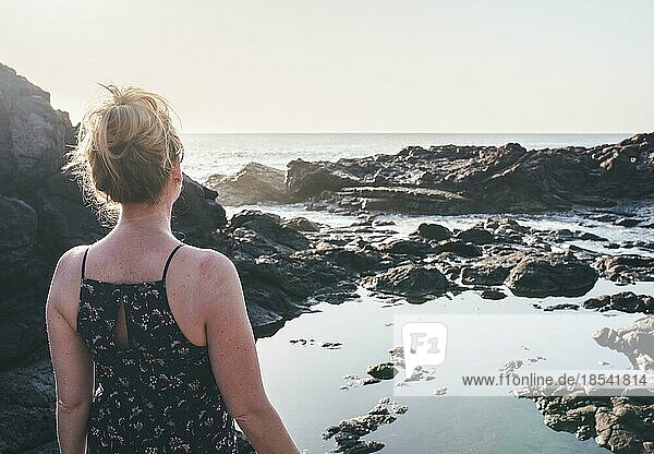 Blonde Frau im Sommerkleid steht am felsigen Ufer und schaut auf das Meer im Sonnenlicht