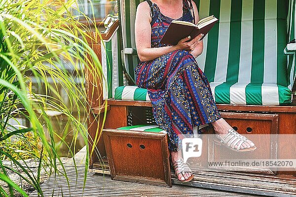 Frau im Sommerkleid entspannt im Strandkorb auf der Terrasse und liest ein Buch