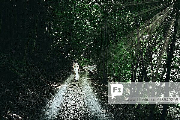Blonde kaukasische Frau im Sommerkleid geht auf schmalem Schotterweg durch dunklen Wald