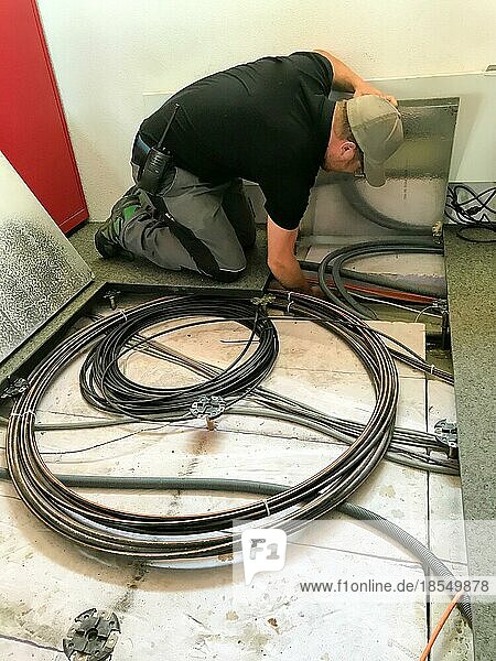 Junger männlicher Elektriker  der an Kabeln im Unterboden arbeitet und Bodenplatten für den Zugang entfernt
