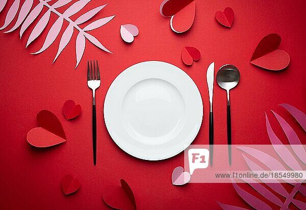 Romantisches Abendessen im Restaurant am Valentinstag  Papierkunst. Weiß leeren Teller  Löffel  Gabel  Messer auf rotem Hintergrund mit Papier Herzen und tropischen Blättern. Valentinstag Datum Konzept Handwerk Origami