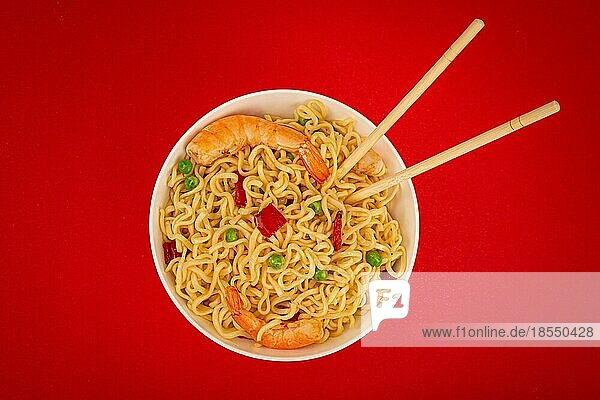 Asiatische chinesische Nudeln in der Draufsicht mit Garnelen und Gemüse in einer weißen Schüssel mit Holzstäbchen auf rotem Papierhintergrund  minimales Foodphoto-Konzept