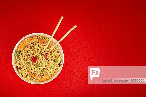 Asiatische chinesische Nudeln in der Draufsicht mit Garnelen und Gemüse in weißer Schüssel mit Holzstäbchen auf rotem minimalem Papierhintergrund  minimales Foodphoto Konzept  Platz für Text
