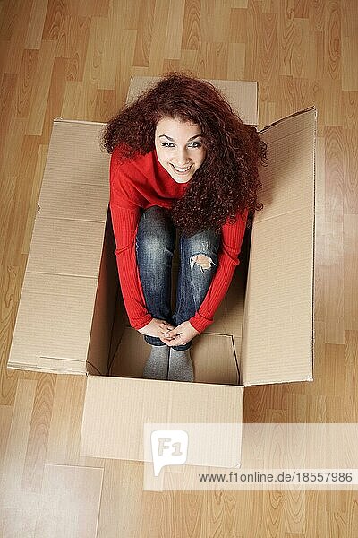 Lächelnde junge Frau sitzt im Karton in ihrem neuen Zuhause