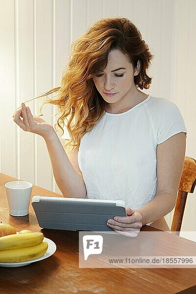 Junge Frau benutzt einen Tablet-Computer  während sie zu Hause am Küchentisch Kaffee trinkt